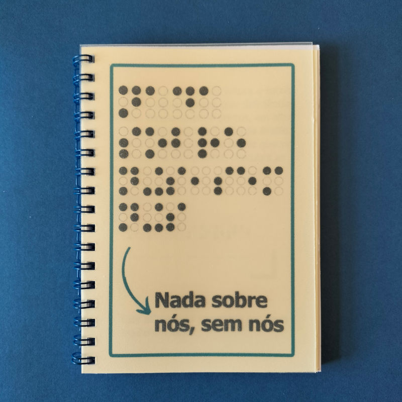 Caderno ACAPO - Alfabeto Braille. Na capa, a frase "Nada sobre nós, sem nós" escrita com os caracteres Braille em relevo, e a tinta, também com relevo.