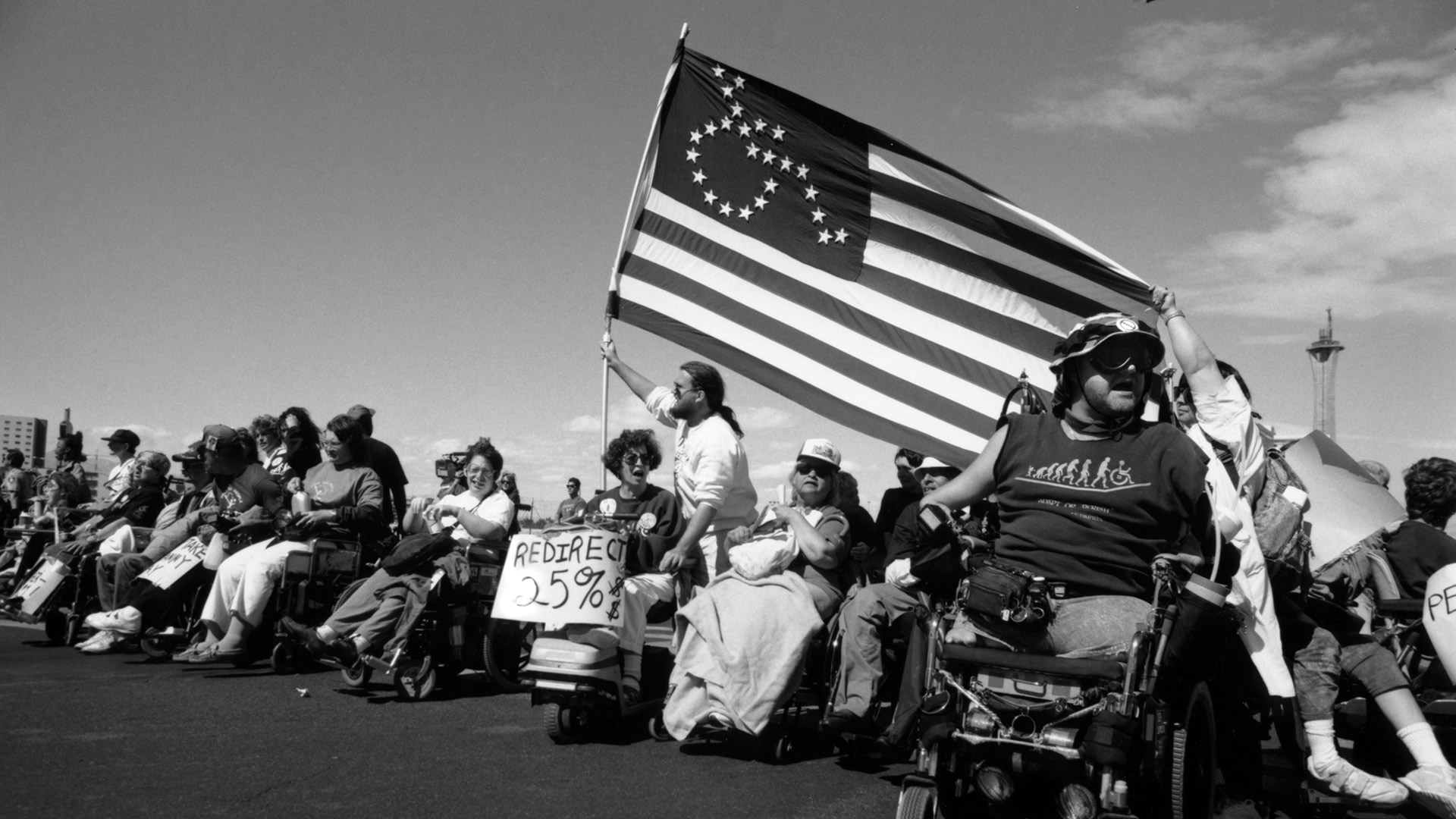 Fotografia, a preto de branco, de pessoas em cadeira de rodas numa manifestação. Uma das pessoas transporta a bandeira dos Estados Unidos, onde no lugar das 50 estrelas está o pictograma da deficiência motora. 