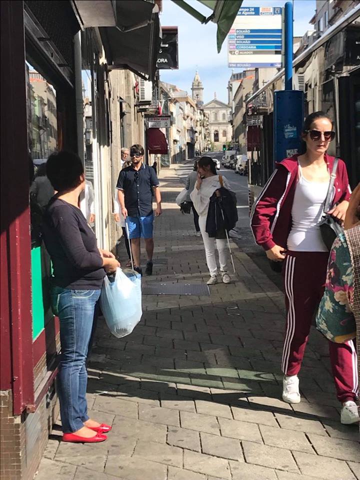 Pessoa de bengala e olhos vendados a descer rua sob o olhar de duas pessoas. 