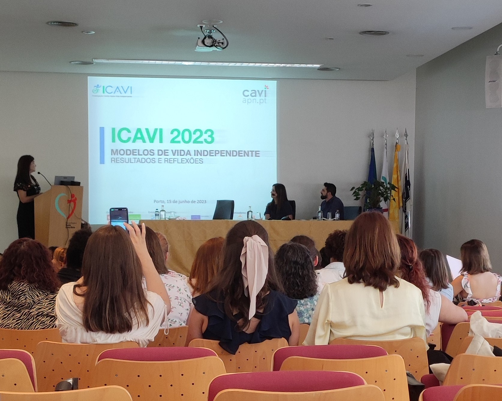 Fotografia tirada durante a conferência onde é possível ver-se, de costas, as pessoas sentadas a assistir e, ao fundo, a mesa de oradores e a projeção da apresentação.