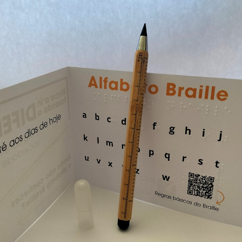 Lápis Infinito 3 Funções ACAPO de pé, encostado ao postal que o acompanha e que contém o alfabeto Braille em tinta e Braille.
