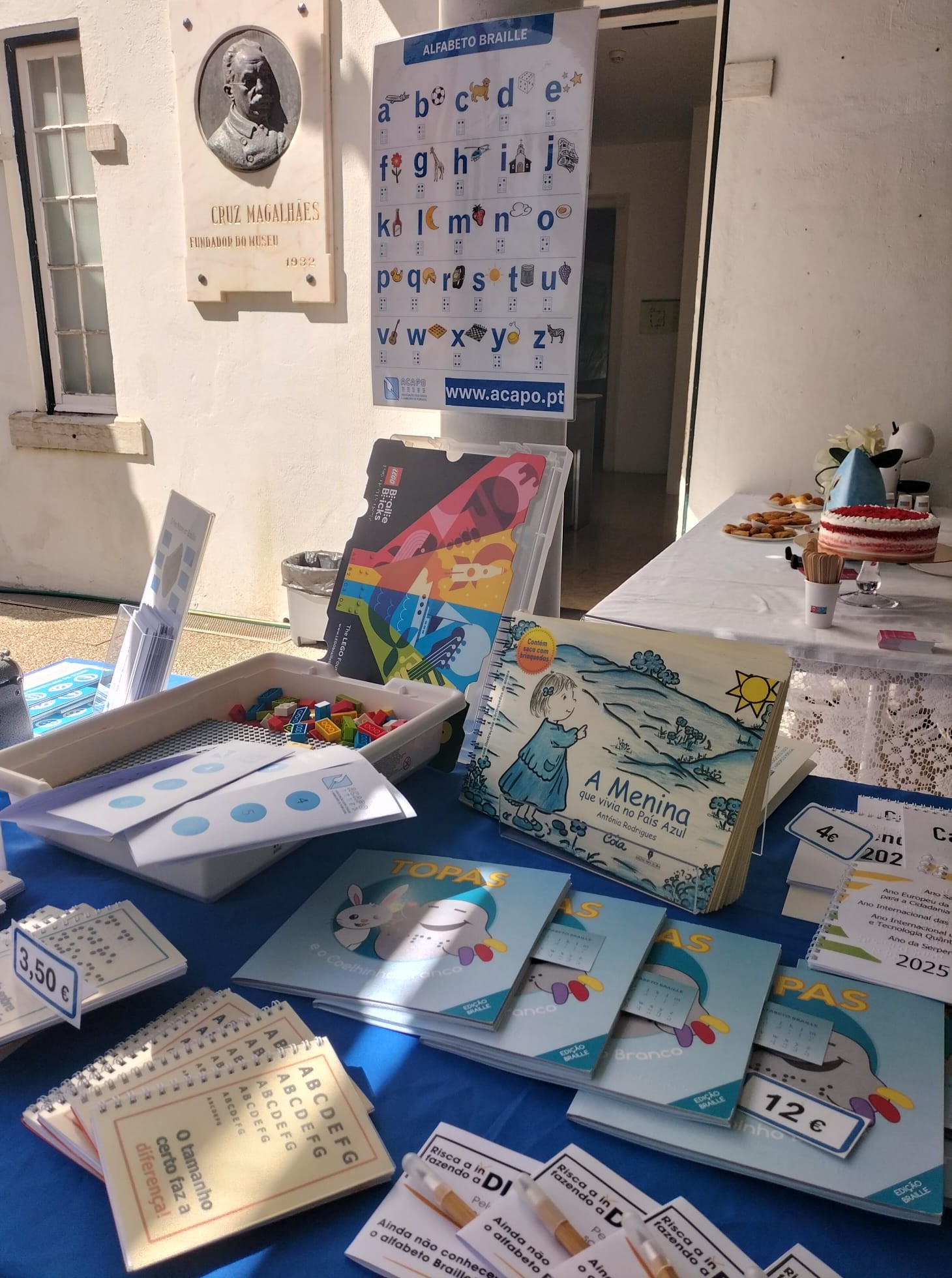 Banca com artigos para venda, cartazes de divulgação do alfabeto Braille, artigos de demonstração como o LEGO Braille Bricks e o livro A menina que vivia no país azul. Na imagem vê-se também uma pedra mármore com o busto do fundador do museu.