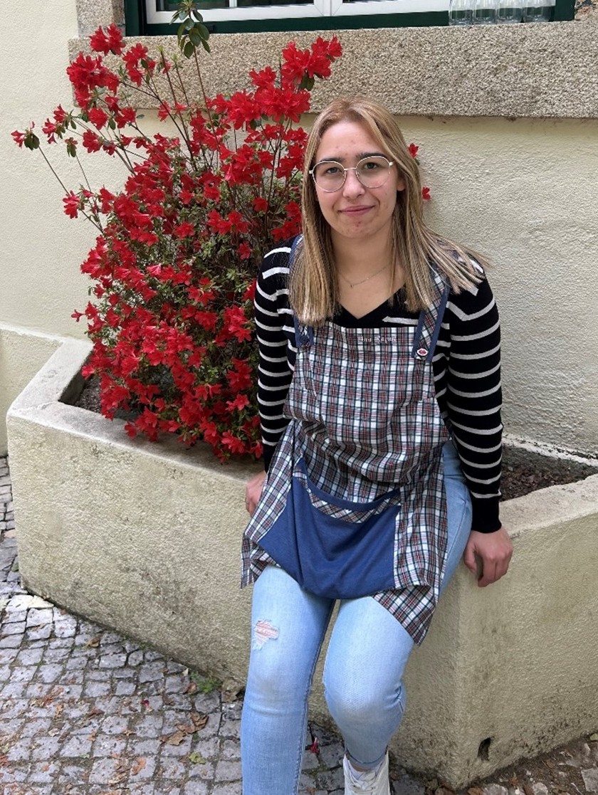 Raquel Oliveira, sorridente, pousa para a fotografia sentada ao lado de um arbusto florido.