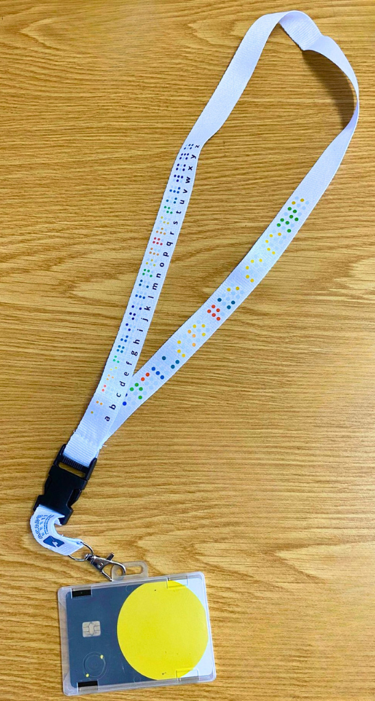 Fita de pescoço Alfabeto Braille Fita de pescoço Alfabeto Braille em cima de uma mesa. A fita contém um gancho metálico onde está colocada uma bolsa transparente com um passe de transportes públicos.