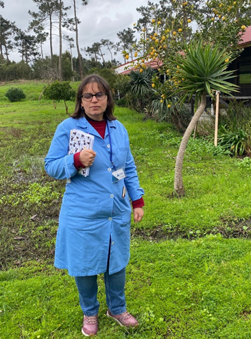 Fotografia de Isabel, ao ar livre, num jardim. Isabel está de pé, veste uma bata azul e segura na mão um caderno.