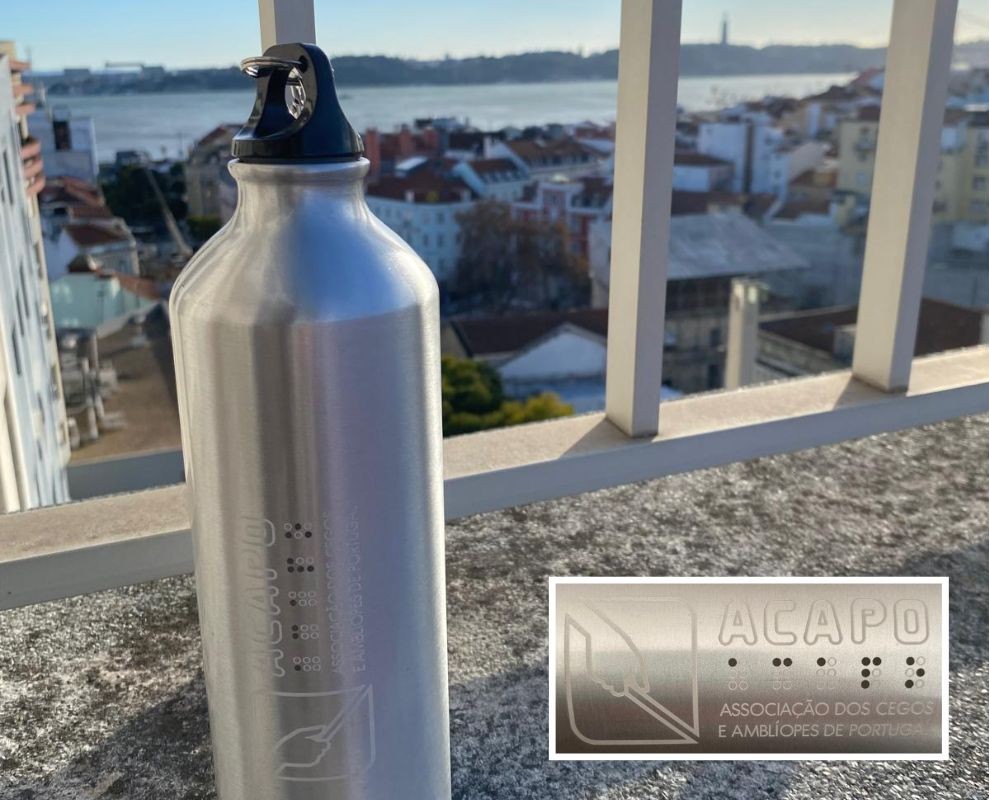 Garrafa de Desporto ACAPO Garrafa de Desporto ACAPO num parapeito vendo-se a vista sobre a cidade de Lisboa, nomeadamente sobre o Rio Tejo. Ao longe, vê-se o Cristo Rei. No canto inferior direito, imagem aproximada destacando o logotipo da ACAPO impresso na garrafa.