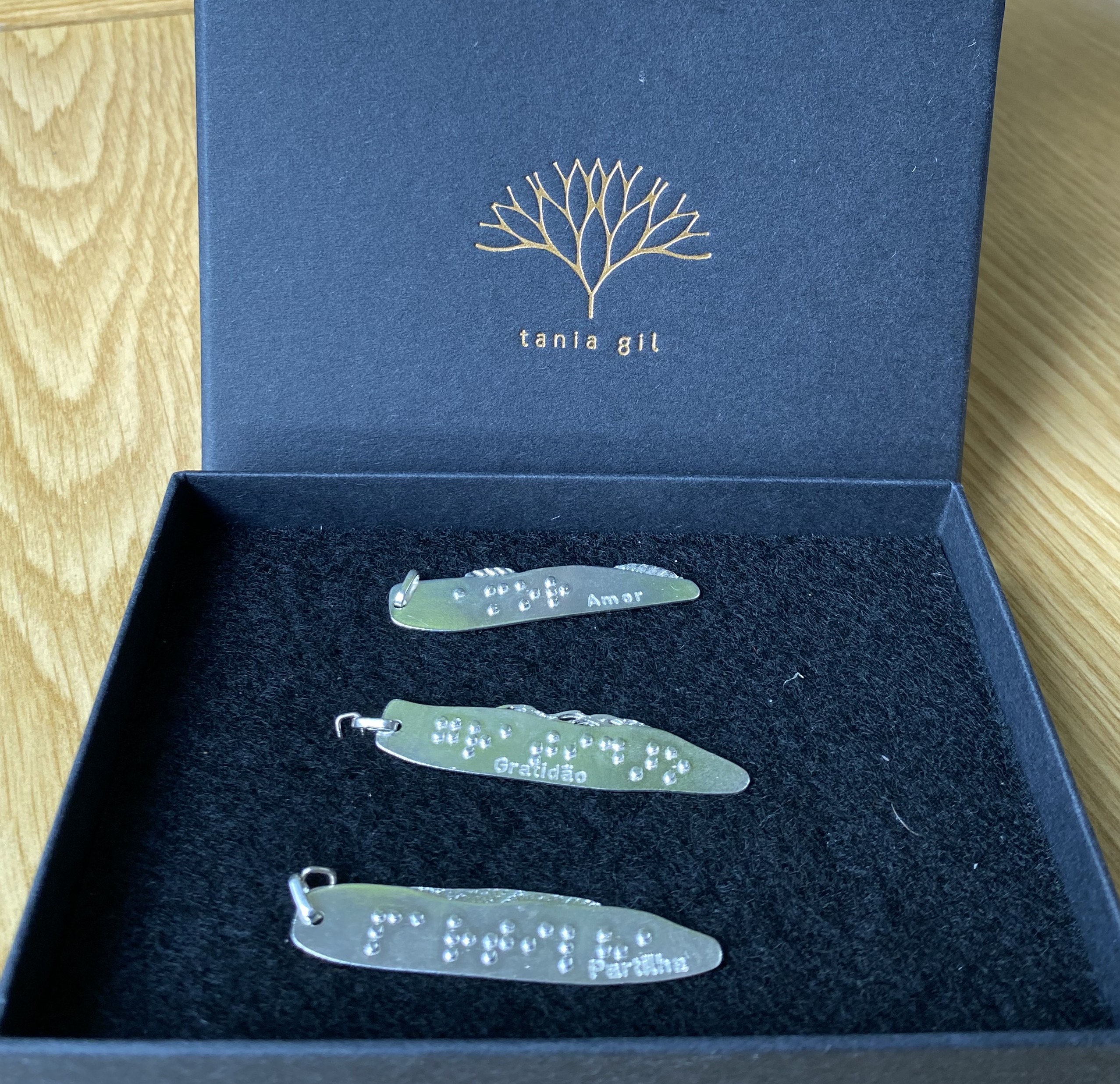 Em cima de uma mesa está uma caixa azul de cartão aberta com os três pendentes no interior. De cima para baixo, vemos as peças Amor, Gratidão e Partilha. Vemos também a tampa da caixa com o logotipo da Tânia Gil.