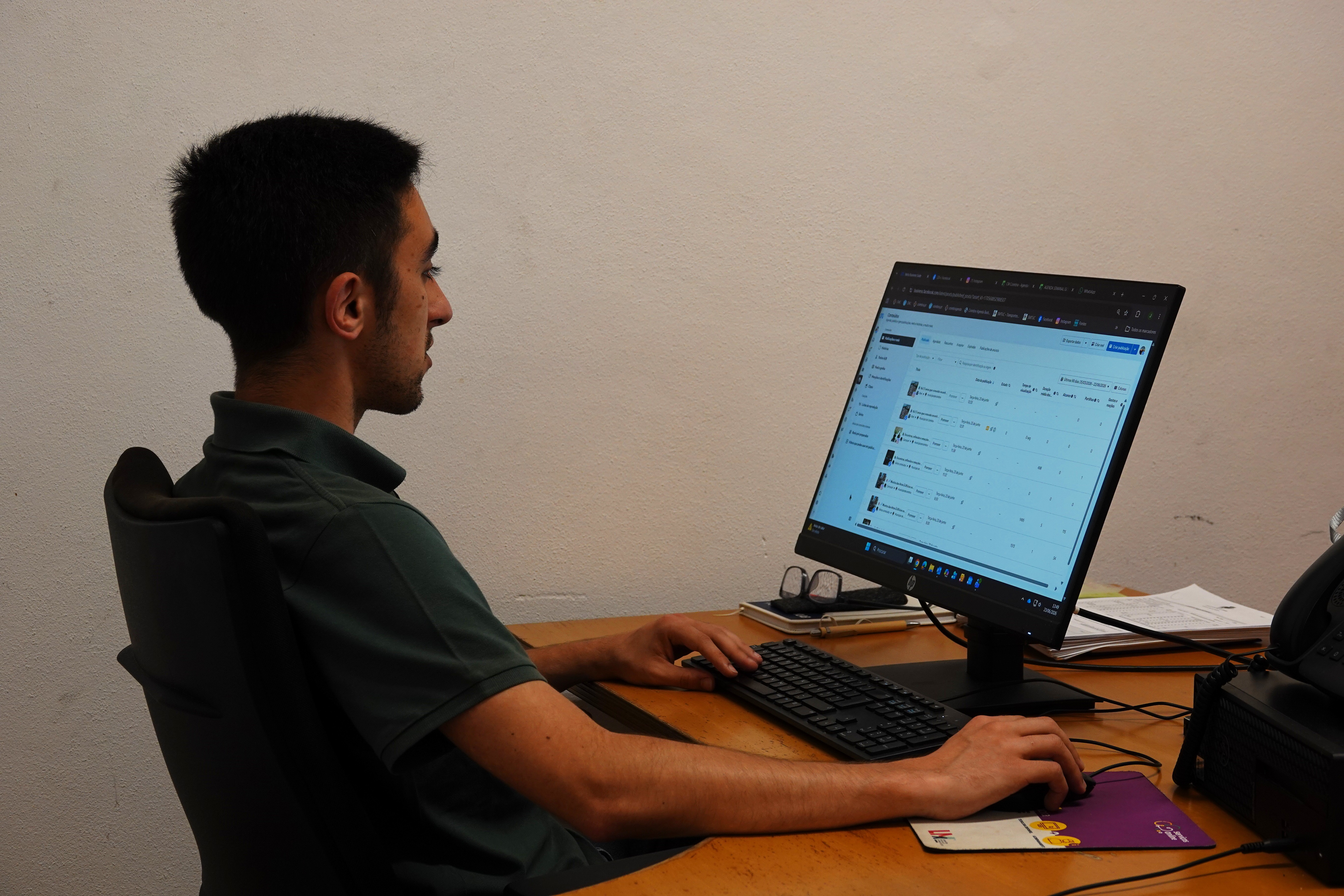 João Sobral Fotografia de João Sobral, sentado, enquanto trabalha ao computador. Em cima da mesa, para além do monitor, teclado e rato que está a utilizar, vemos também algumas folhas, um caderno, uma caneta e uns óculos.