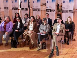 Representantes dos 10 projetos finalistas sentados em cadeiras. Por trás deles, é possível ver-se diversos roll ups com a descrição de cada projeto.