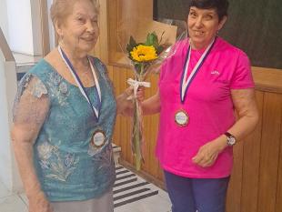Uma das participantes pousa sorridente para a fotografia com uma medalha de participação ao peito. A senhora segura também uma gerbera que lhe está a ser entregue por Aciolinda Coelho, Secretária da Direção da Delegação de Lisboa. 