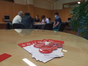 Folheto e crachá da iniciativa Biblioteca Humana de Valongo, vendo-se ao fundo, desfocado, o grupo de participantes a conversar em redor de uma mesa redonda.