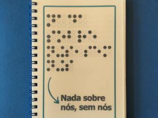 Caderno ACAPO - Alfabeto Braille. Na capa, a frase "Nada sobre nós, sem nós" escrita com os caracteres Braille em relevo, e a tinta, também com relevo.