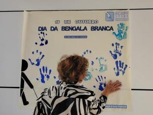 Uma senhora a “carimbar” a sua mão num painel decorado com várias marcas de mãos em tinta azul. No topo, o título 15 de outubro – Dia da Bengala Branca, seguido pelo slogan “De mãos dadas com a inclusão” e o logótipo da ACAPO.