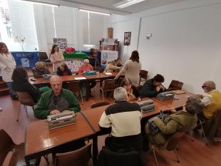 Grupo de participantes, sentados, durante o workshop de Braille criativo. À sua frente têm uma máquina Braille.