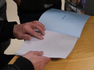 Uma mão a ler o livro "Topas e o Coelhinho Branco", aberto, na página onde tem início a história em Braille