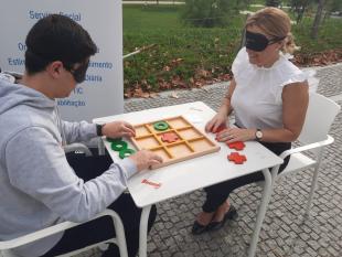 Dois participantes, um rapaz e uma senhora, sentados a uma mesa, de olhos vendados enquanto jogam ao jogo do galo em relevo.