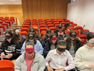 Participantes sentados, de olhos vendados, num auditório, durante a sessão de informação. Nas mãos seguram o alfabeto Braille, distribuído pelas técnicas, e alguns dos alunos exploram-no através do toque.