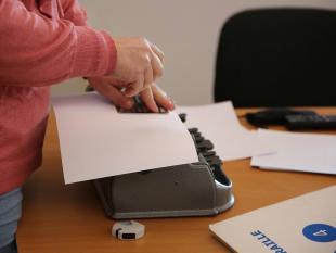 Fotografia ampliada onde se vê as mãos da nossa Técnica de Braille enquanto utiliza a punção e a régua Braille para escrever.