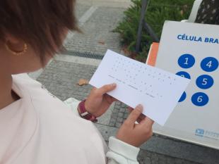 Rapariga com um alfabeto Braille nas mãos. Na fotografia é possível ver-se também uma célula Braille impressa numa folha A4, que se encontra em cima da mesa.