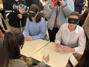 Participantes, de olhos vendados, sentados à mesa. Em cima da mesa estão copos de vidro que os alunos exploram e reconhecem através do toque.