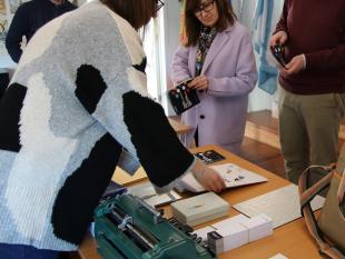 2 participantes leem atentamente um dos nossos folhetos sobre deficiência visual, em frente à mesa onde estão expostos diversos materiais relacionados com a temática, nomeadamente: máquina Braille, cartaz com a célula Braille impressa e alfabetos Braille.