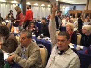 Vista geral da sala em que se encontram sentados os delegados. Primeiro plano da imagem encontra-se Tomé Coelho e Rodrigo Santos que está com braço direito no ar e na mão segura um envelope. 