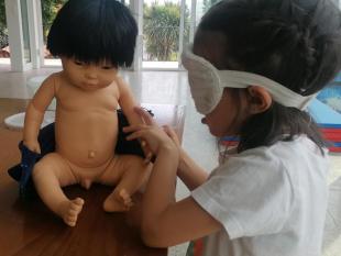Menina, de olhos vendados, a limpar com uma toalha o seu boneco, após lhe ter dado banho.
