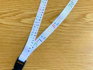 Fita de pescoço Alfabeto Braille em cima de uma mesa. A fita contém um gancho metálico onde está colocada uma bolsa transparente com um passe de transportes públicos.