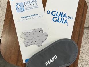 Sobre uma mesa, um folheto informativo da Delegação de Vila Real, um folheto informativo "O Guia do Guia" e uma venda de olhos com a palavra ACAPO 