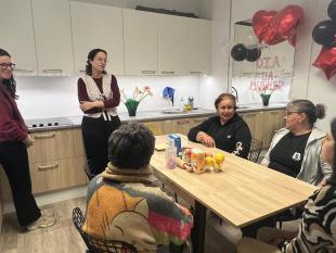 Na cozinha da Delegação as participantes conversam entre si antes da atividade. Em cima da mesa, veem-se os produtos e utensílios que serão utilizados. A cozinha está decorada com balões e num espelho está escrito Dia da Mulher.