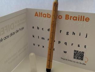 Lápis Infinito 3 Funções ACAPO de pé, encostado ao postal que o acompanha e que contém o alfabeto Braille em tinta e Braille.
