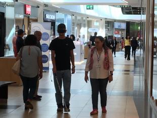 Um rapaz caminha pelo LeiriaShopping de olhos vendados e a utilizar uma bengala branca. Ao seu lado, caminha uma das técnicas para o apoiar.