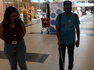 Um rapaz caminha pelo LeiriaShopping de olhos vendados e a utilizar uma bengala branca. Ao seu lado, caminha uma das técnicas para o apoiar.