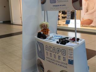 Banca e roll up da ACAPO no LeiriaShopping. Em cima da banca estão diversos artigos relacionados com a sensibilização para a deficiência visual.