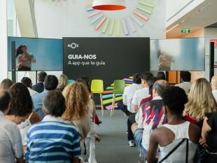 Fotografia tirada do fundo da sala vendo-se as pessoas, de costas, sentadas em cadeiras. Ao fundo está uma tela, com o logo da NOS e a frase "Guia-NOS A app que te guia". Ao lado da tela estão dois ecrãs onde está a ser projetada uma imagem de uma mulher.