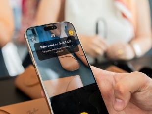 Fotografia aproximada vendo-se um telemóvel nas mãos de uma pessoa e por trás caixas de óculos Ray-Ban. No ecrã do telemóvel vemos aberta a App Guia-NOS. Em segundo plano, em modo desfocado, vemos uma senhora com uma bengala branca na mão. 