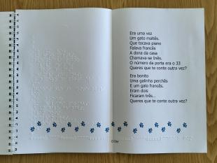Livro aberto numa das lengalengas vendo-se o texto em caracteres ampliados e em Braille e, por baixo, o desenho das pegadas deixadas pelas patas de um gato.
