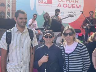 Junto ao palco, três participantes pousam sorridentes para a foto com os elementos da banda HMB que estão no palco.