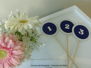 Em cima de uma mesa, um ramo de flores e as placas para votação de 1, 2 ou 3 pontos. 