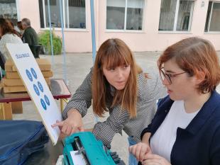 Uma das visitantes tenta escrever na máquina Braille enquanto olha atentamente para uma célula braille impressa a tinta, em tamanho gigante, e ouve as indicações de uma das nossas técnicas.