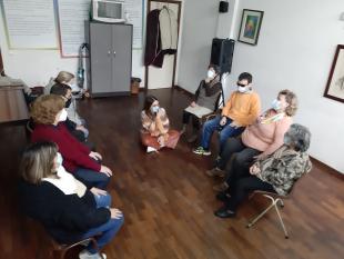 Participantes sentados numa cadeira, ouvem atentamente as explicações da terapeuta que se encontra sentada no chão com as pernas cruzadas
