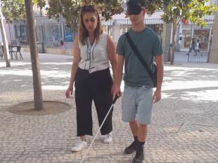Um rapaz caminha de olhos vendados e a utilizar uma bengala branca. Ao seu lado, está uma das técnicas para o apoiar.