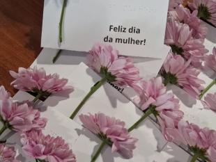 Em cima de uma mesa, várias gerberas cor-de-rosa com um postal e tinta e braille onde está escrito Feliz dia da mulher!
